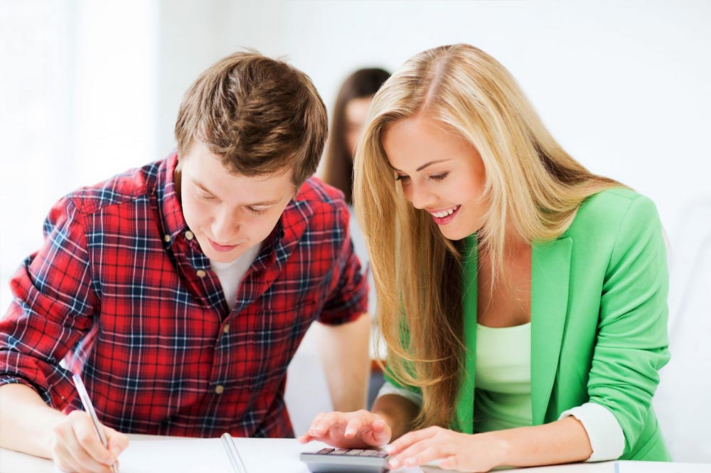 A teenage boy and girl sat at a desk studying. The boy is writing while the girl is typing on a calculator.