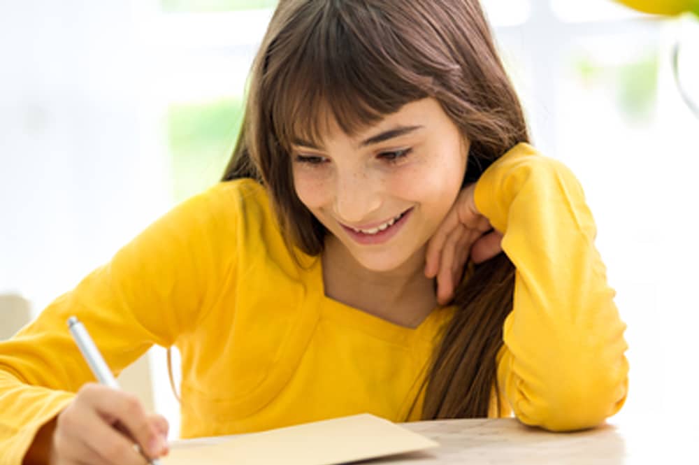 A young girl sat at a table doing schoolwork. She is smiling.